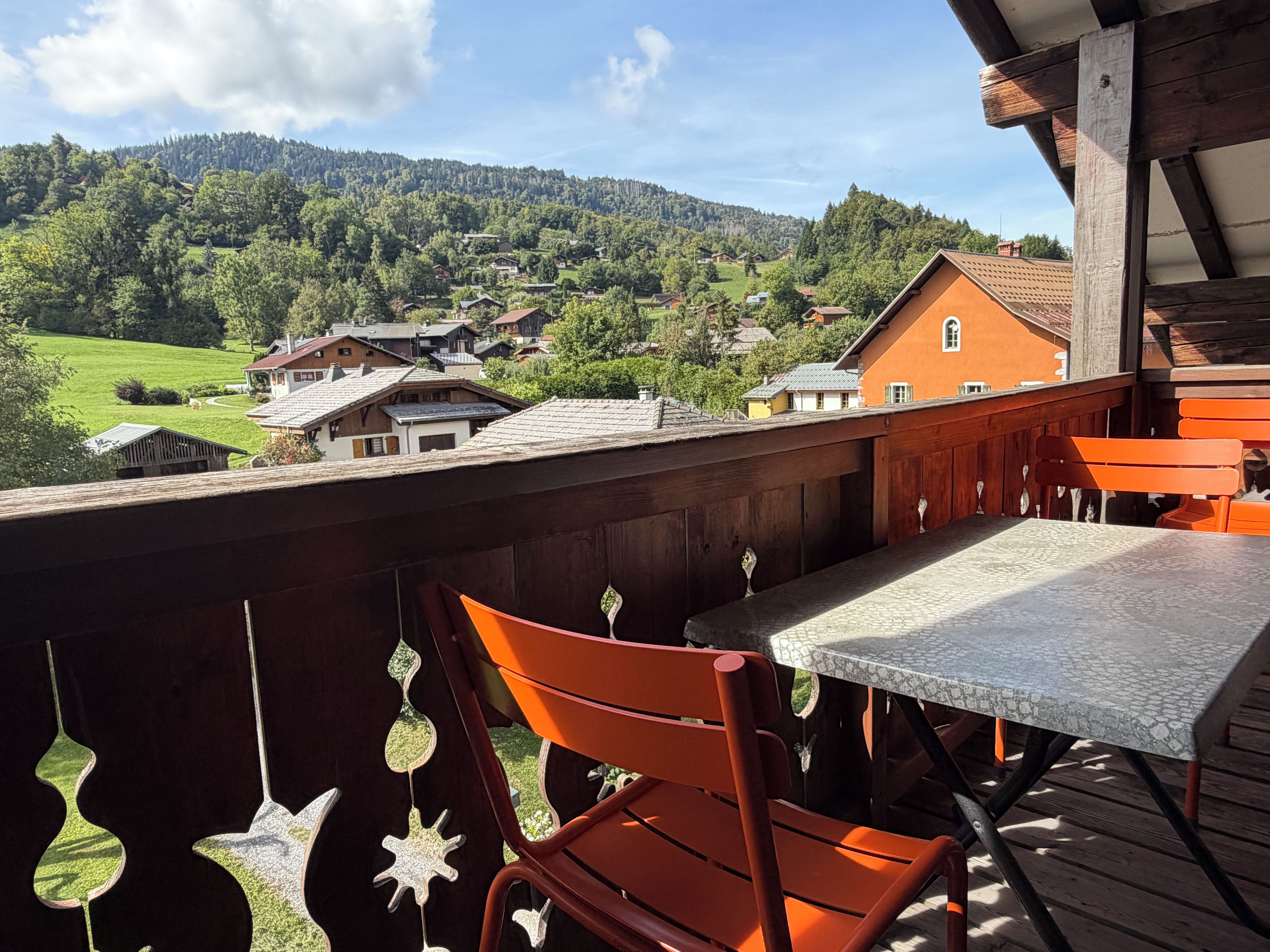 A wooden deck with a table and chairs, overlooking a village with houses and a forested hill.
