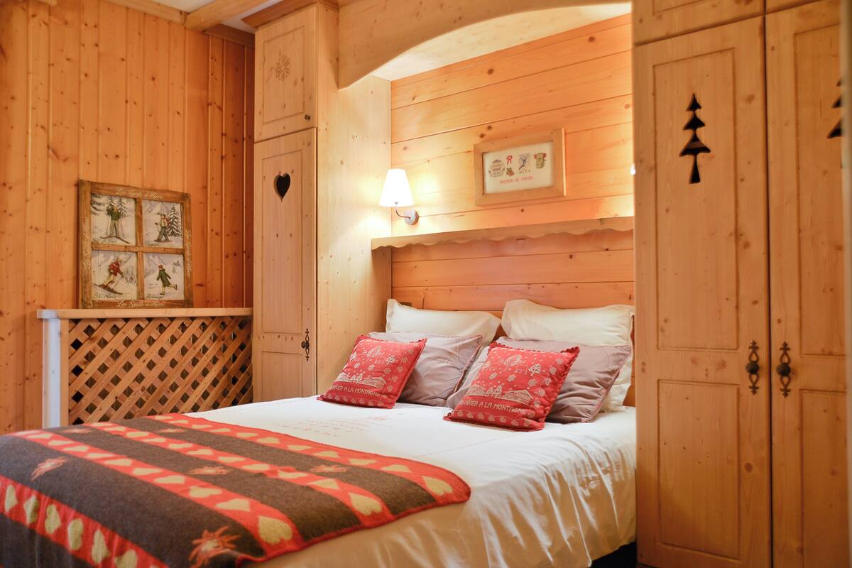 A wooden bedroom with a bed, pillows, a quilt, a wall-mounted lamp, and a framed picture.
