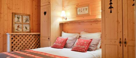 A wooden bedroom with a bed, pillows, a quilt, a wall-mounted lamp, and a framed picture.