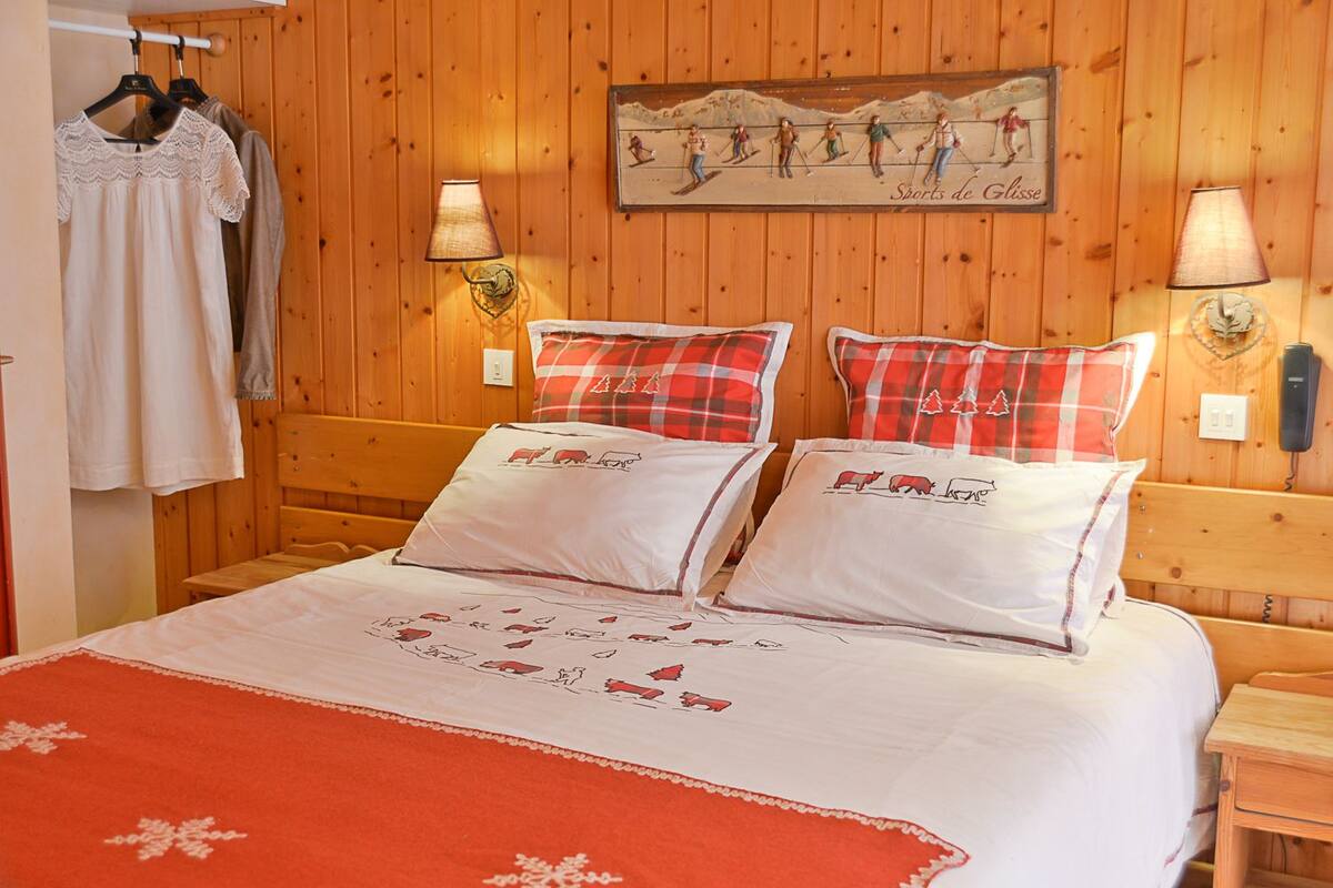 A wooden-paneled room with a bed featuring red and white bedding, a painting of skiers, and a lamp on the wall.