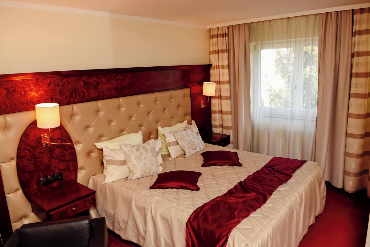 A hotel room with a large bed, two bedside tables, a headboard, and a window with curtains.