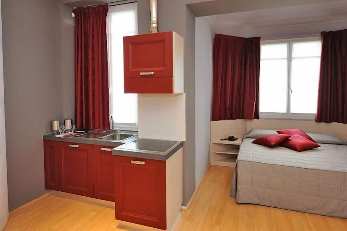 A compact room with a kitchenette, a bed, and a window with curtains.