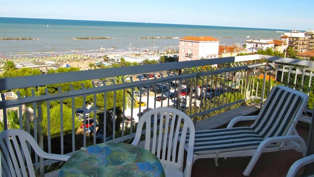 Varanda com cadeiras e mesa brancas, vista para uma marina com barcos e uma praia.