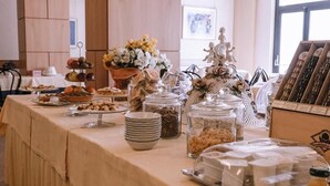 Mesa de buffet com diversos alimentos, incluindo pastelaria, frutas e cereais, coberta por um pano de mesa bege.