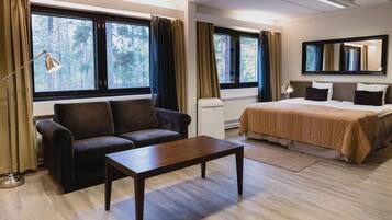 A modern hotel room with a sofa, a bed, a wooden coffee table, and large windows with curtains.