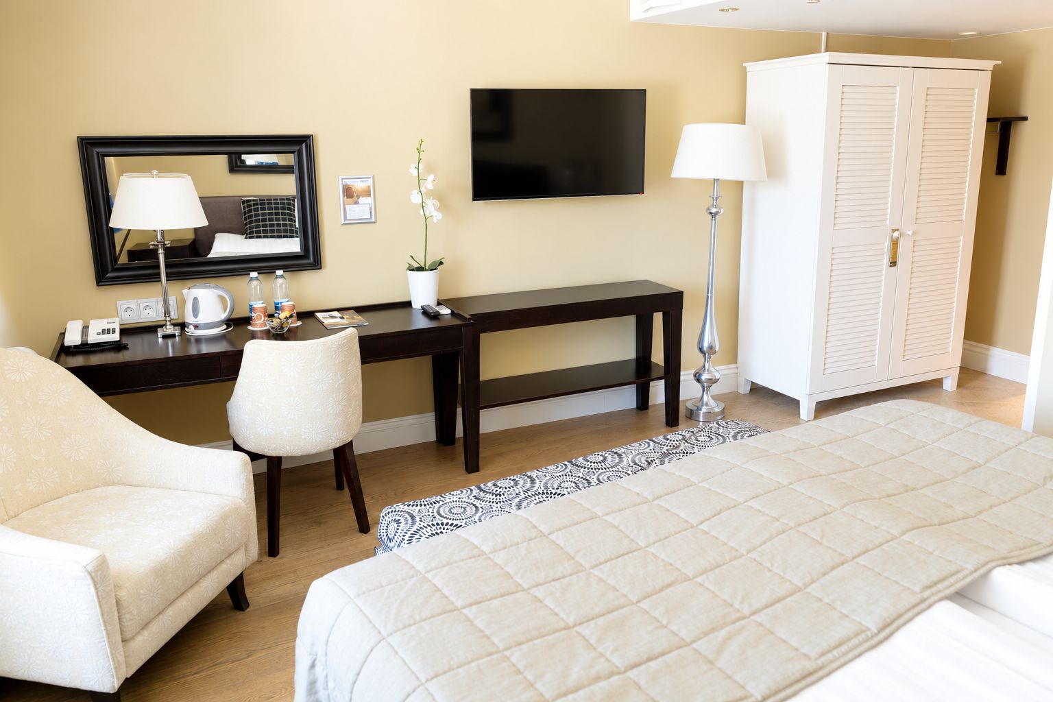 Quarto de hotel com uma cama, escrivaninha com cadeira, televisão, guarda-roupa e mesa de cabeceira com abajur.