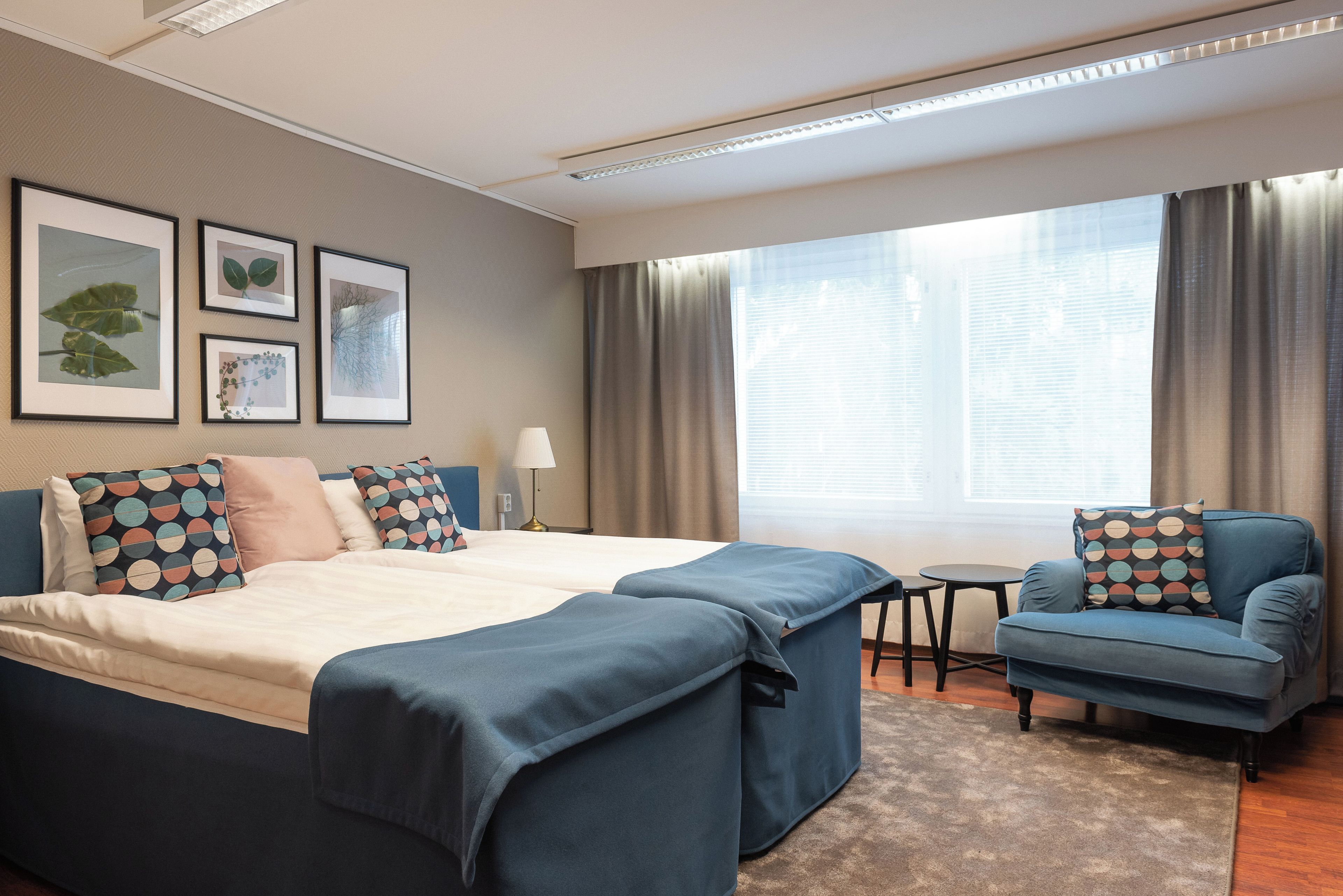 A hotel room with a bed, a chair, a small table, and a window with curtains. There are framed pictures on the wall.