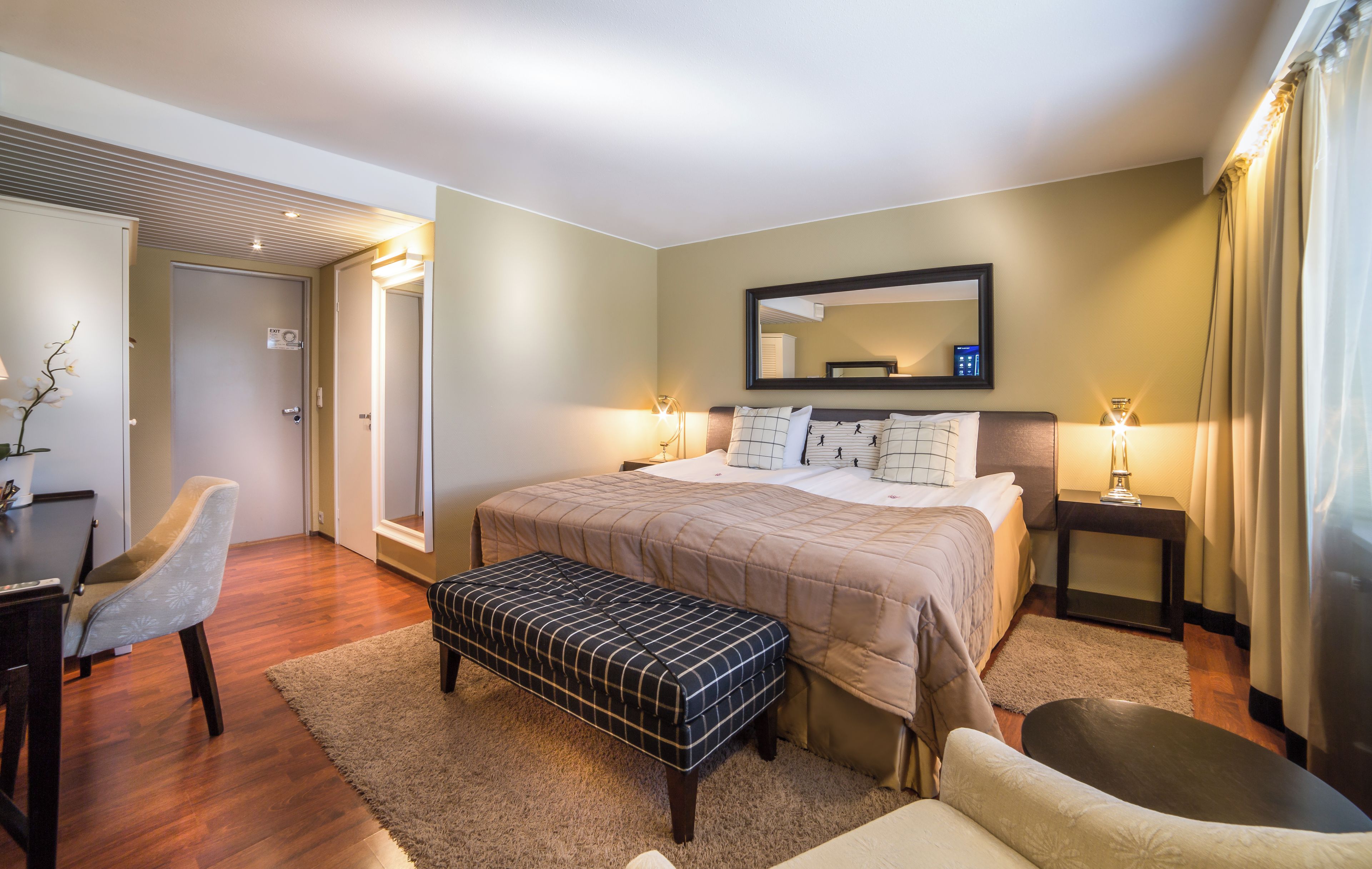 A hotel room with a bed, bedside tables, a desk, a chair, a bench, and a mirror.