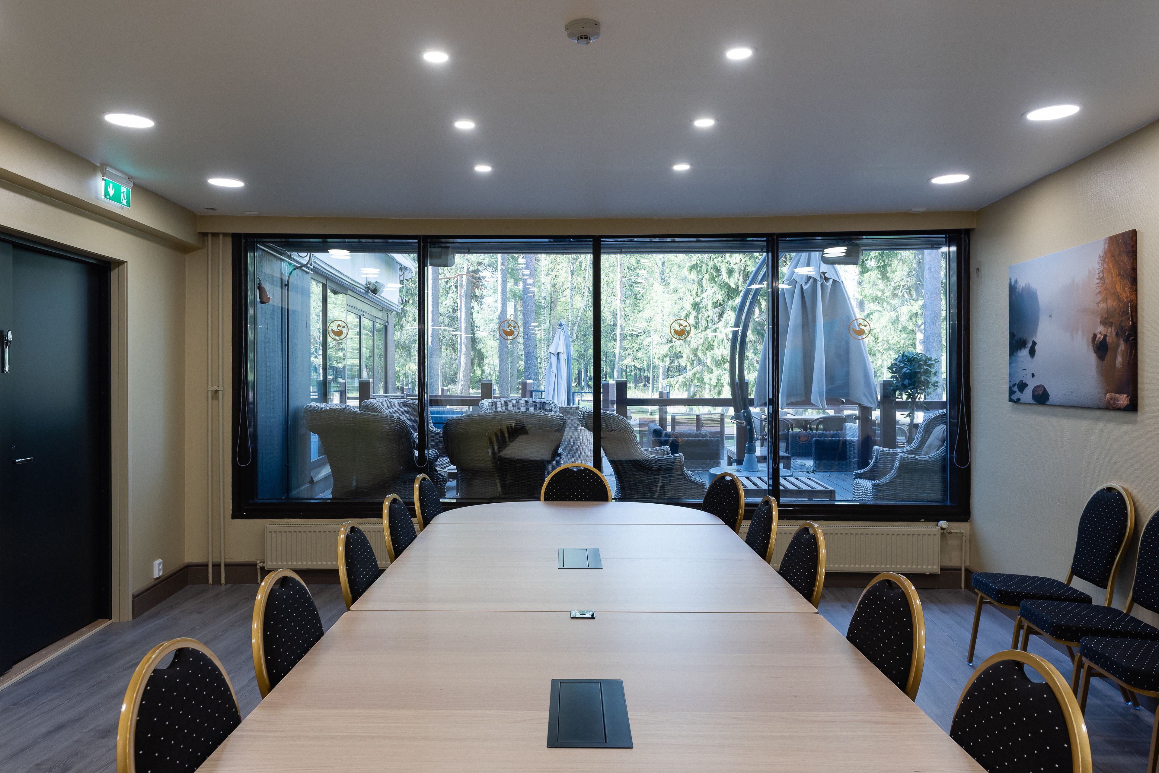 A conference room with a long table, chairs, a projector screen, and large windows with a view of outdoor seating.