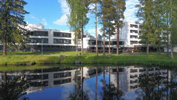 A modern building complex with a reflective water body in front, surrounded by trees and a clear sky.