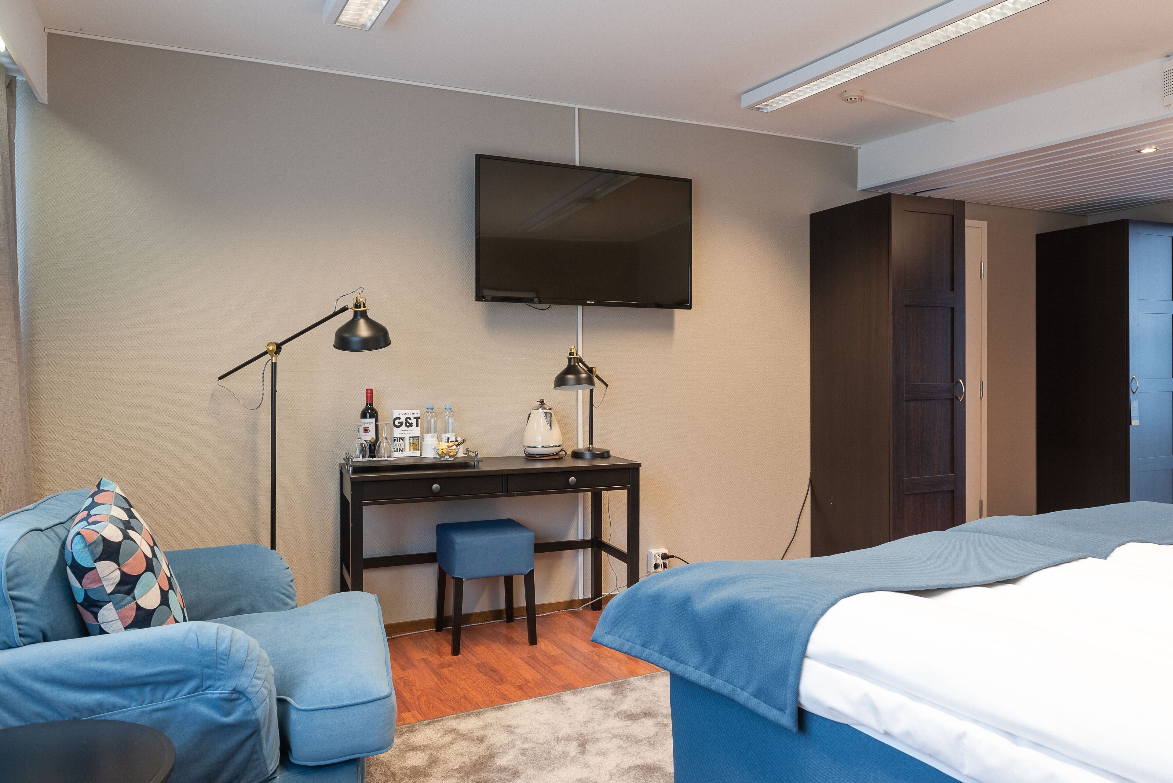 A hotel room with a bed, a desk with a chair, a TV mounted on the wall, and a small table with a lamp.