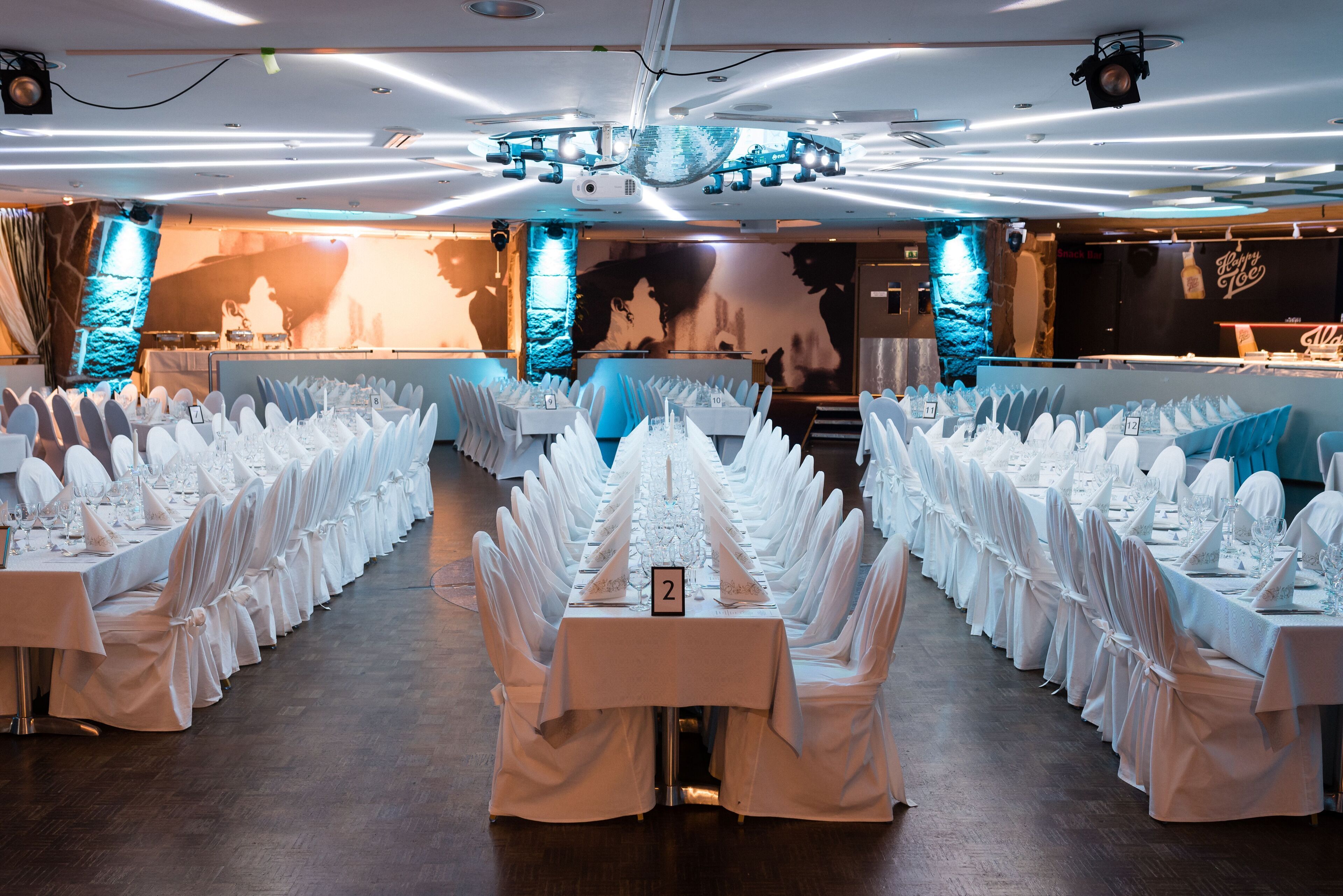 A banquet hall with rows of tables set for a formal event, featuring a central stage and a backdrop with a silhouette.