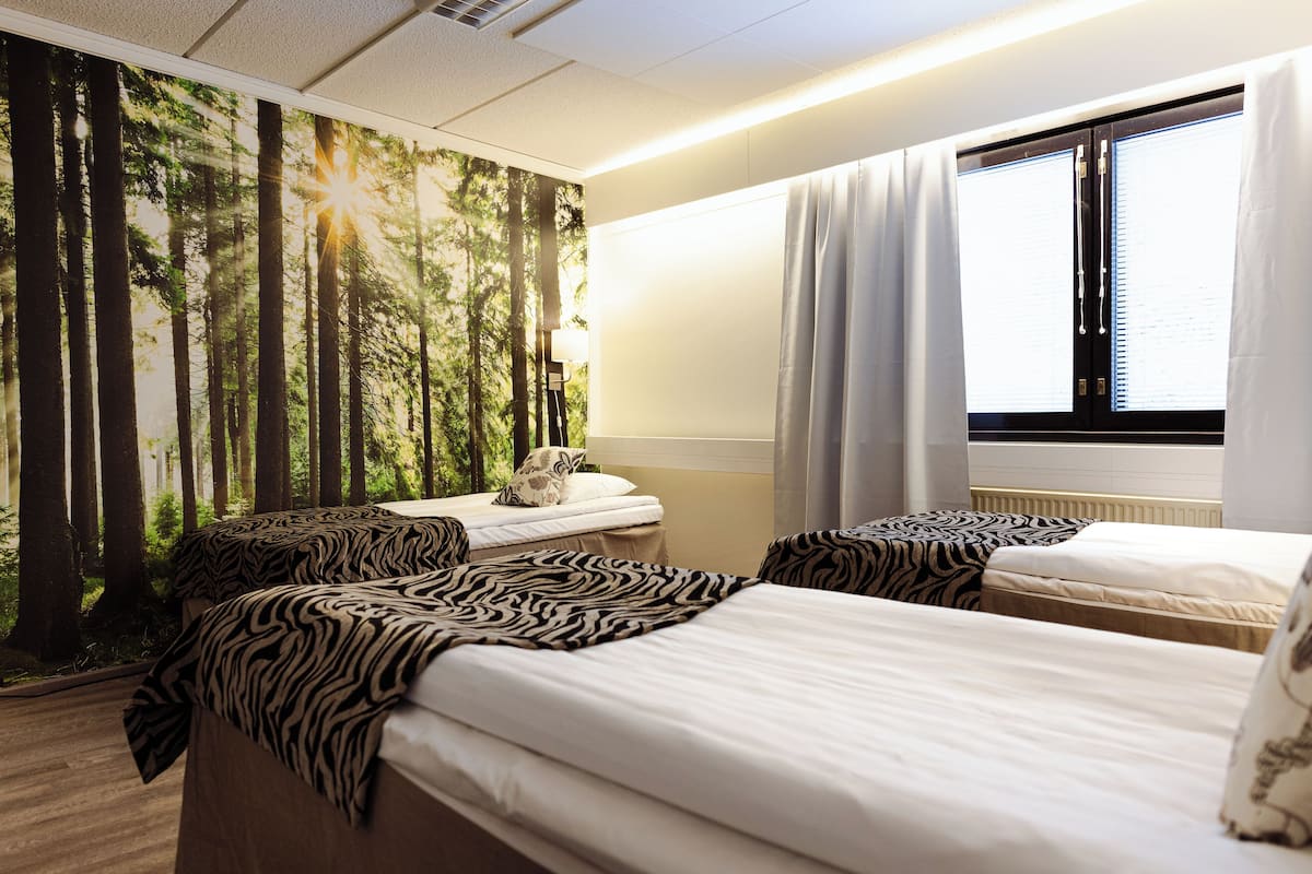 A hotel room with two beds, a forest-themed wall mural, a window with curtains, and a nightstand with a lamp.