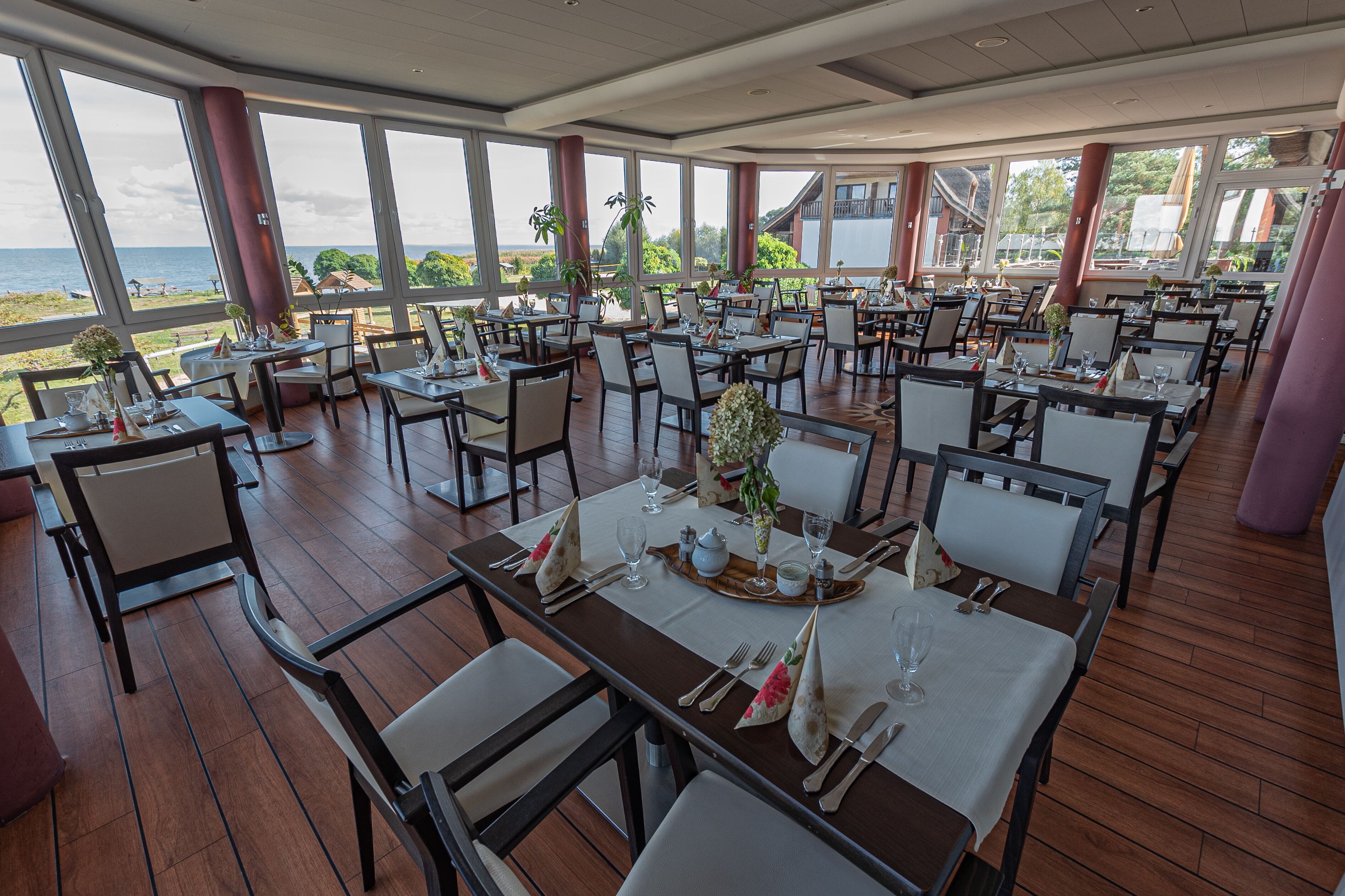 A restaurant with multiple tables set for dining, large windows offering a view of the sea, and indoor plants.