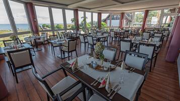 A restaurant with multiple tables set for dining, large windows offering a view of the sea, and indoor plants.