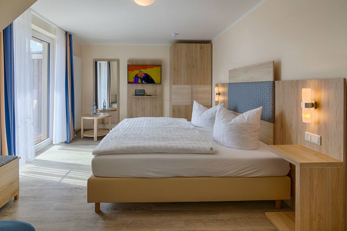 A modern hotel room with a large bed, a small side table, a mirror, and a colorful painting on the wall.