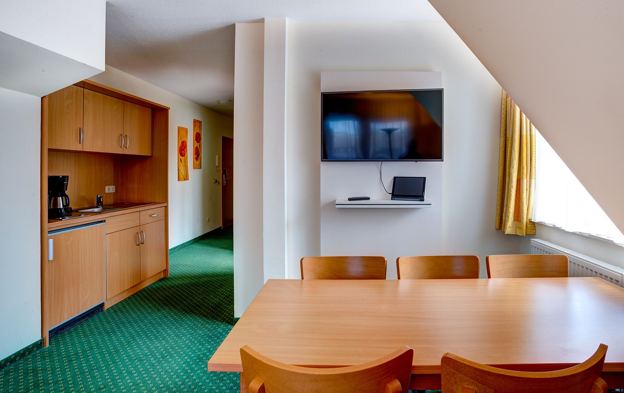 Ein kleines Zimmer mit Esstisch, Holzschränken, einer Küchenzeile, einem Flachbildfernseher und einem Laptop auf einem Regal.