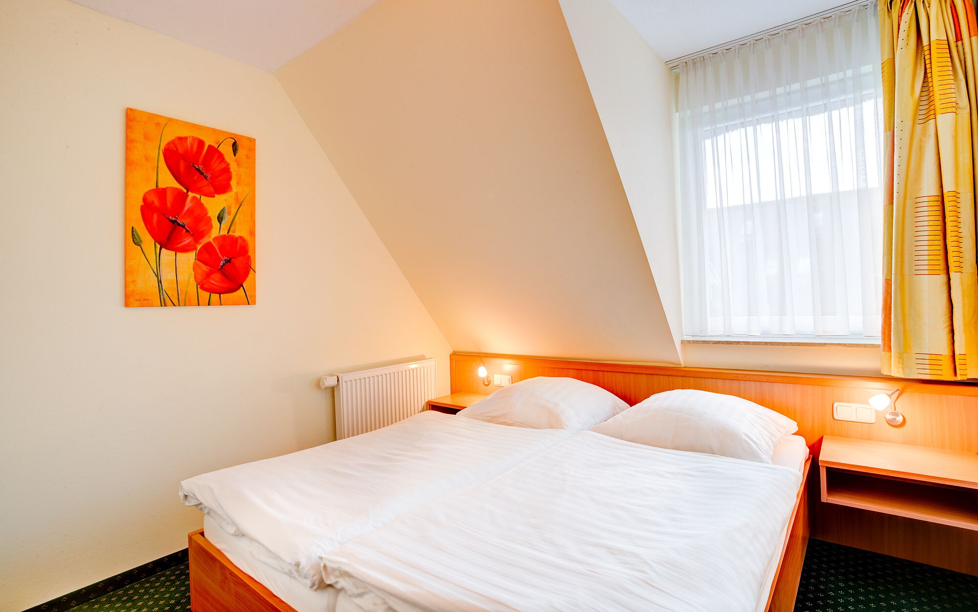 Une chambre d’hôtel avec un lit, un radiateur, un tableau représentant des fleurs rouges et une fenêtre avec des rideaux.