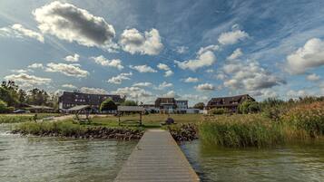 A wooden pier leading to a lakeside property with multiple buildings, a grassy area, and a clear sky with clouds.