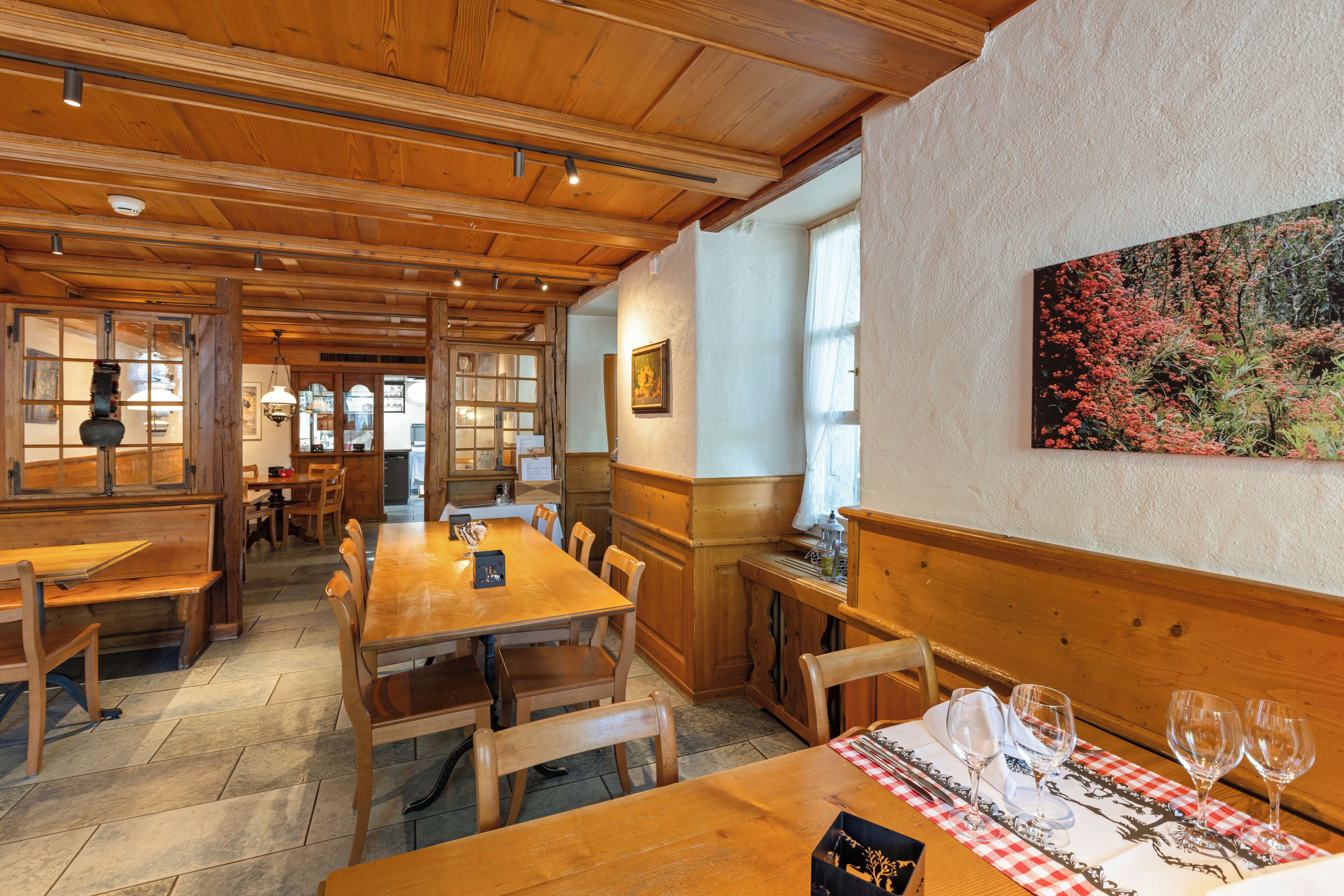 Un espace de repas en bois, avec des tables et des bancs, un tableau accroché au mur et une fenêtre laissant entrer la lumière naturelle.