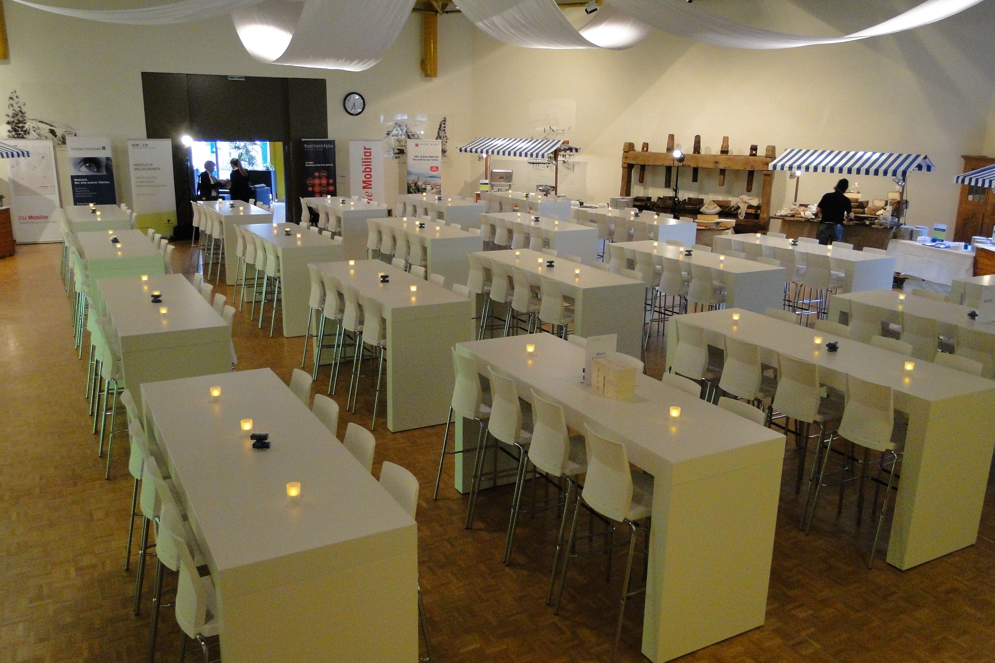 Un grand espace dédié à la salle à manger, avec de nombreuses tables et chaises blanches, un coin cuisine où une personne est en train de préparer un repas, et une horloge accrochée au mur.