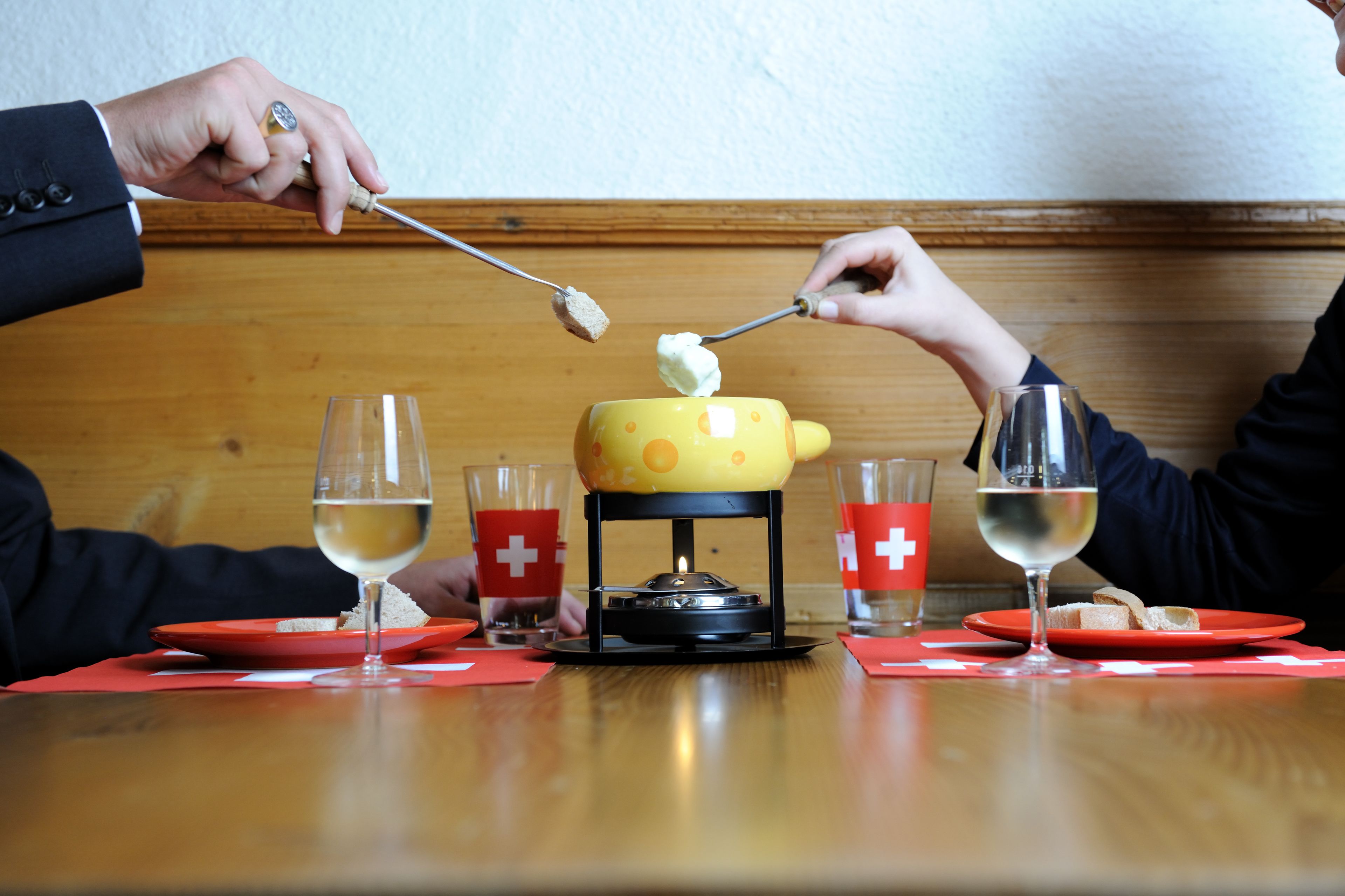 Een eettafel met een kaasfondueset, twee glazen witte wijn en rode servetten met een wit kruis.