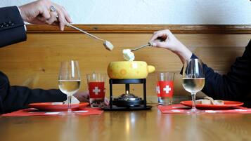 Een eettafel met een kaasfondueset, twee glazen witte wijn en rode servetten met een wit kruis.