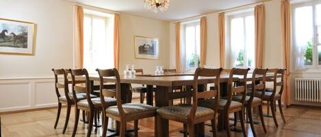 Comedor con una gran mesa de madera y ocho sillas, una araña y ventanas amplias con cortinas.