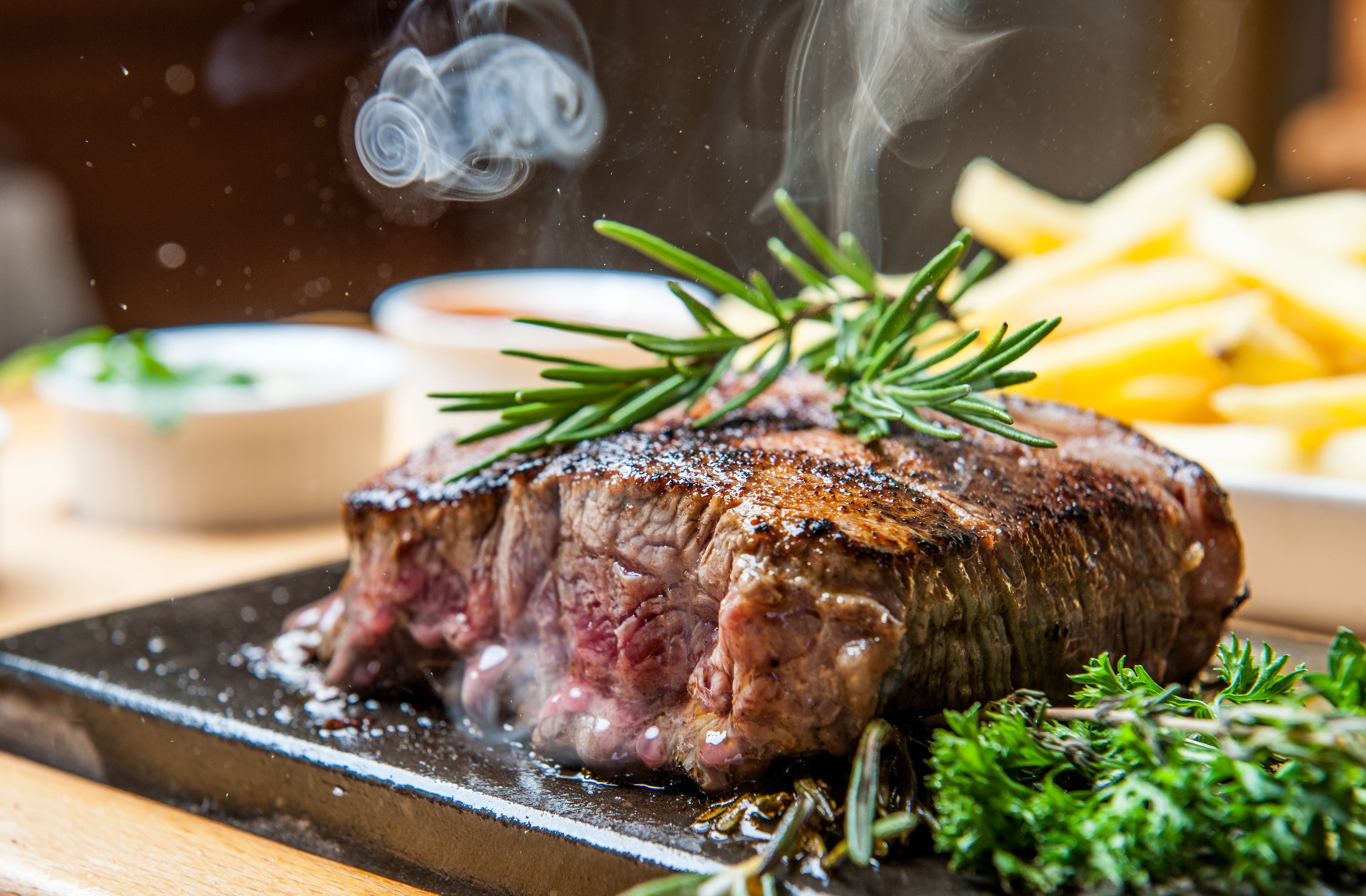 A steak with a garnish of rosemary, accompanied by a side of fries and a small bowl of sauce.
