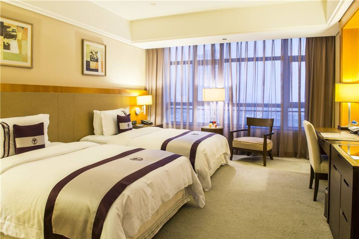 Habitación de hotel con dos camas, un escritorio, una silla y un gran ventanal con cortinas.