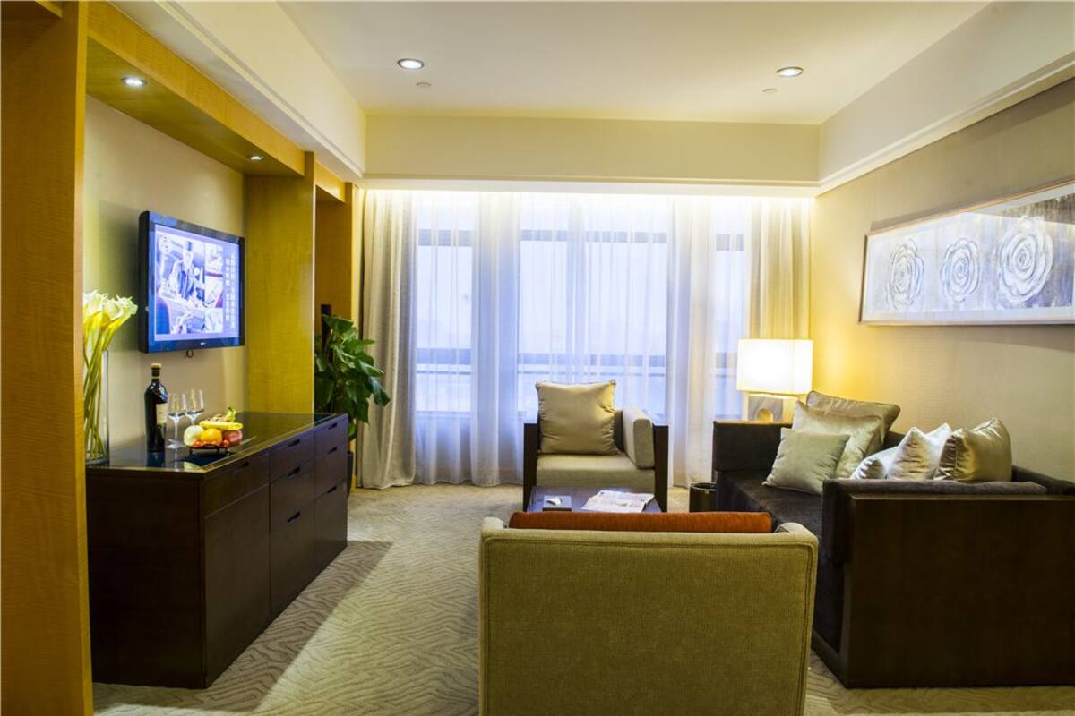 Habitación de hotel con un ventanal grande, un televisor de pantalla plana, un sofá, una mesita de café y un escritorio con una planta y fruta.