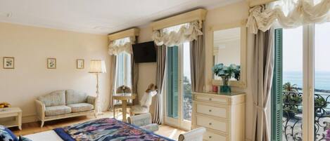 Habitación de hotel con cama, sofá, cómoda, televisor, balcón con vistas y ventana con cortinas.