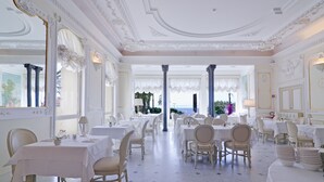 Une salle à manger spacieuse avec des tables et des chaises blanches, de grandes fenêtres ornées de rideaux et une vue sur la mer.