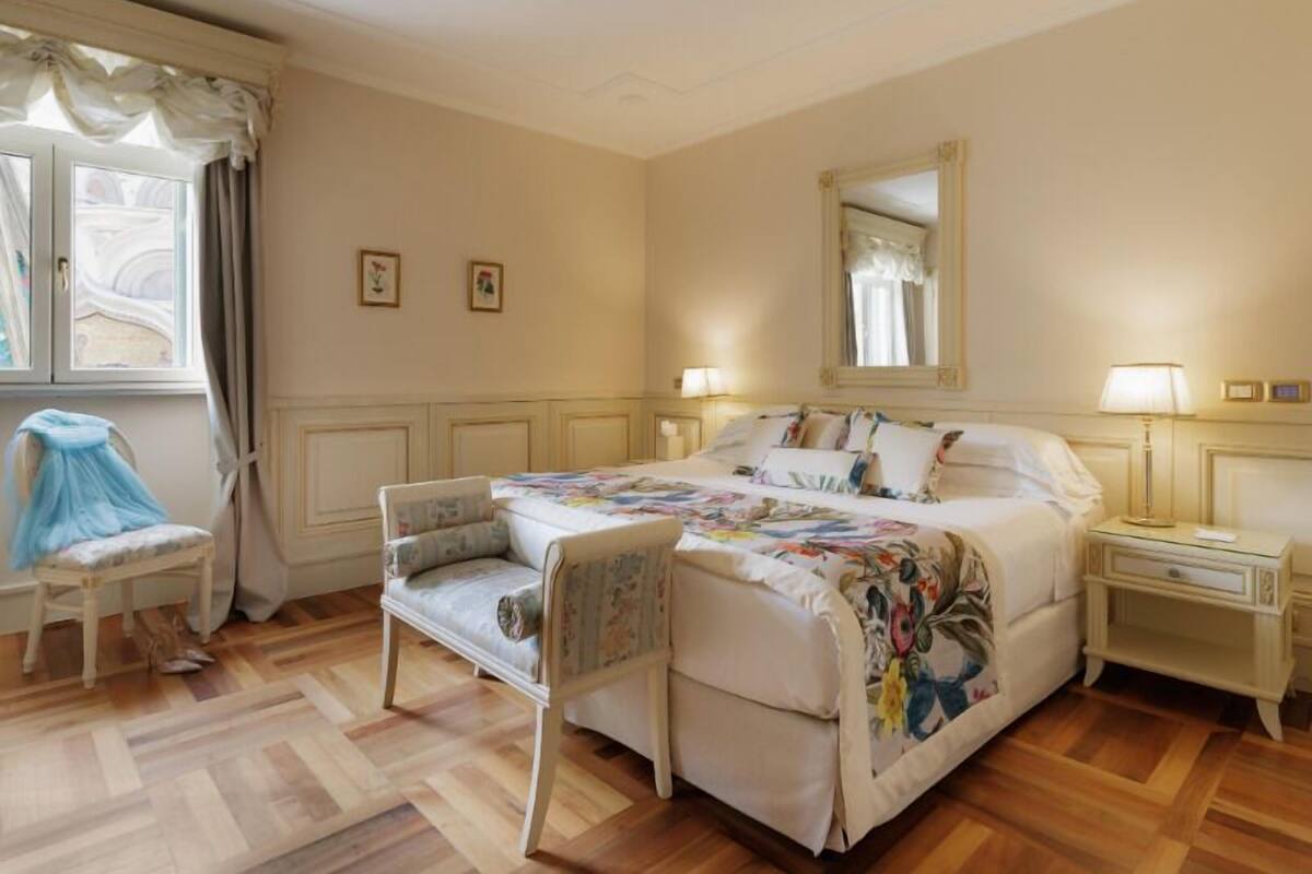 Ein Schlafzimmer mit einem Bett, zwei Nachttischen, einem Sessel, einem Fenster mit Vorhängen und einem Spiegel an der Wand.