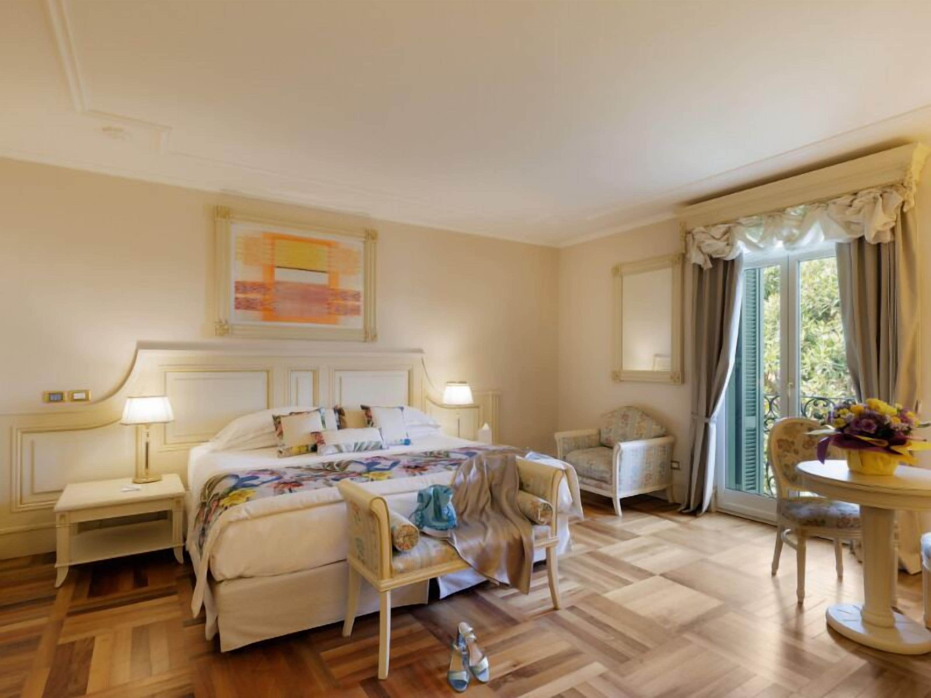 A hotel room with a large bed, bedside tables, a chair, a small table with a vase, and a window with curtains.