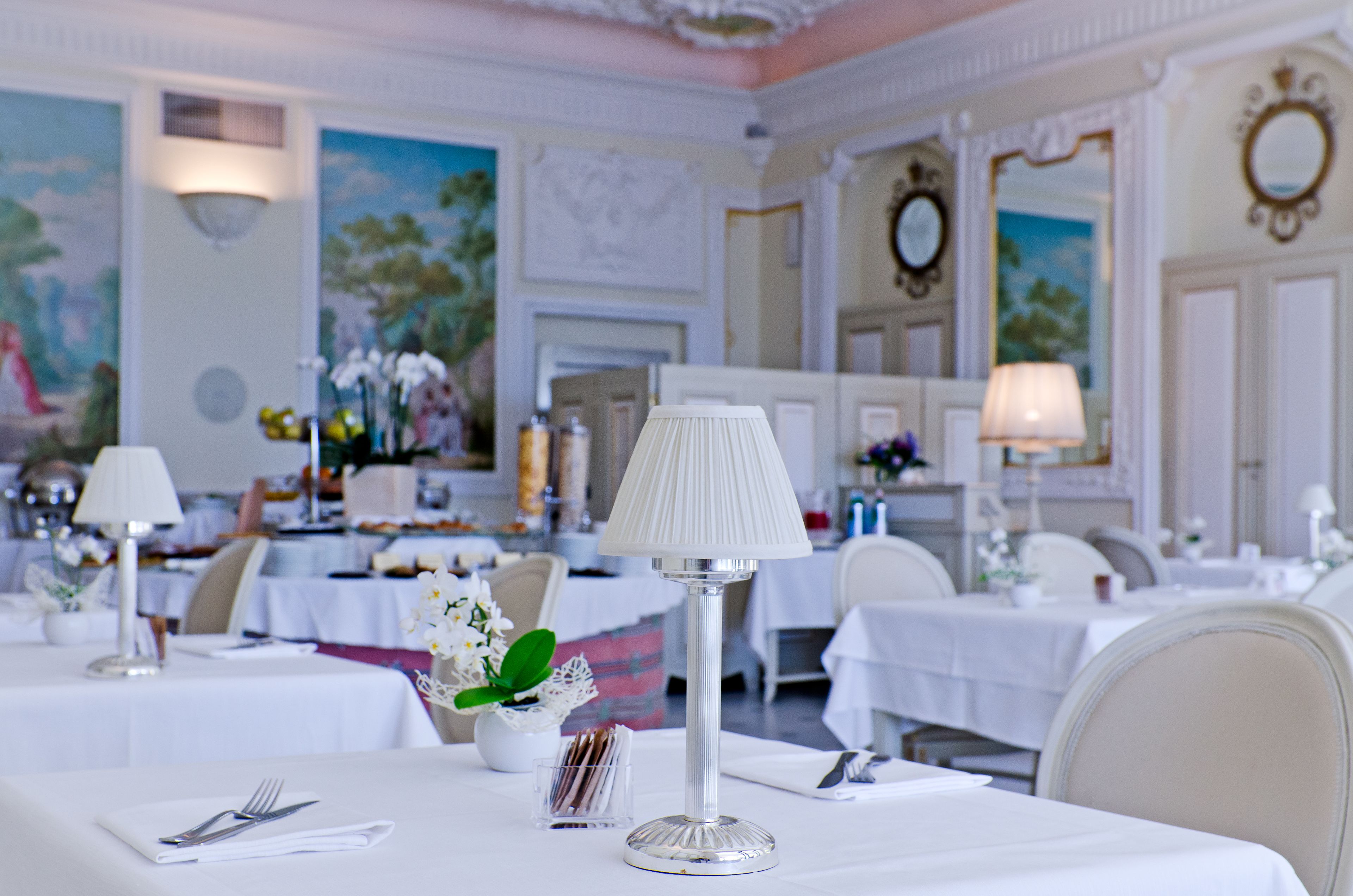 A dining area with tables set for service, white tablecloths, and a central table with a lamp and flowers.