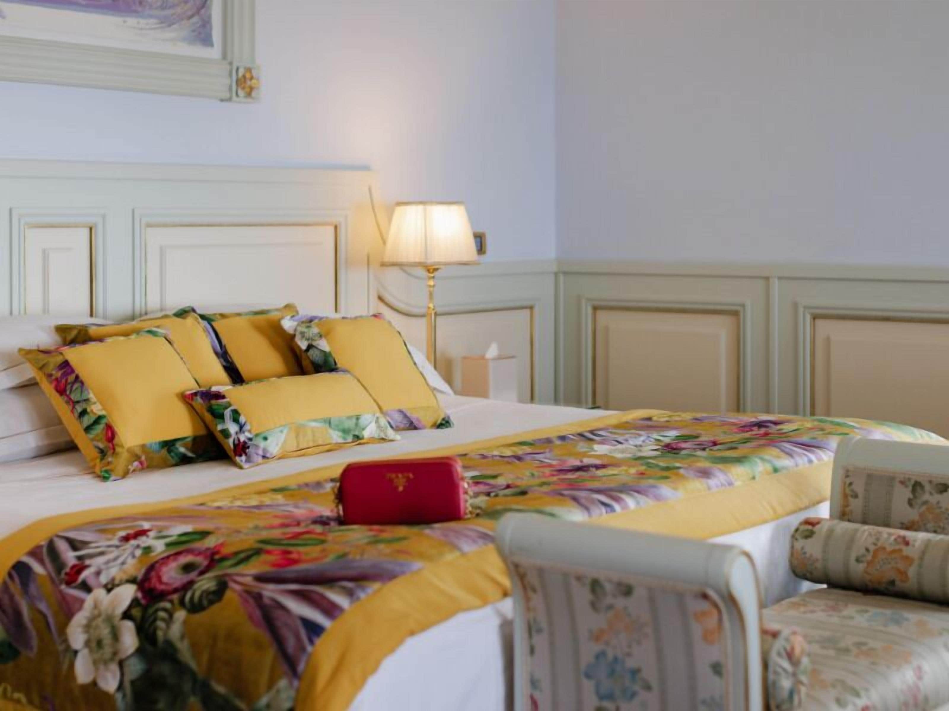 A hotel room with a large bed, a bedside table, a lamp, and a floral-patterned chair.