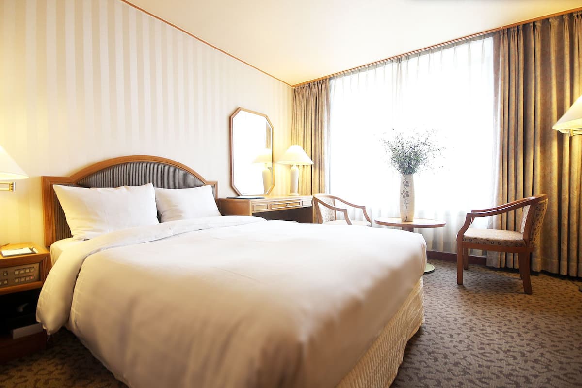 A hotel room with a large bed, a bedside table with a lamp, a chair, a small table with a vase, and a window with curtains.