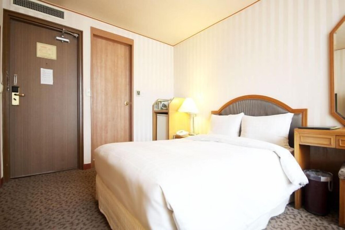 A hotel room with a large bed, a bedside table with a lamp, a wooden headboard, a mirror, and a trash can.