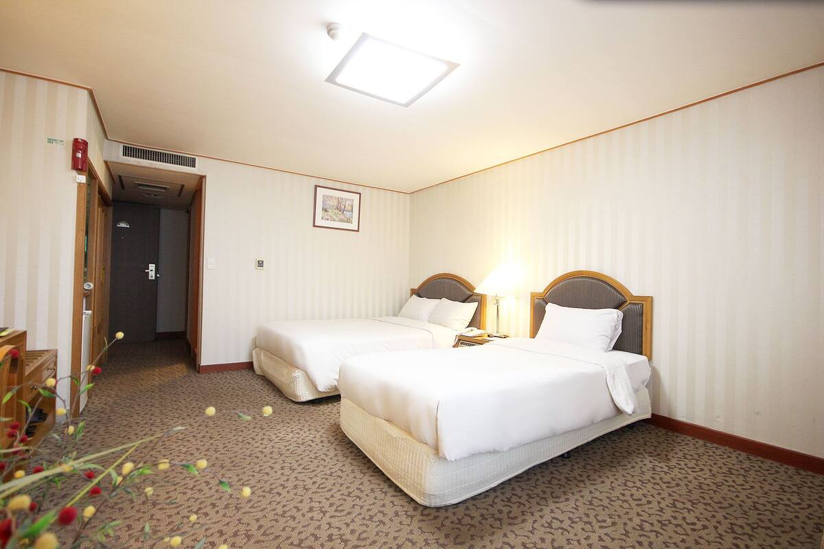 A hotel room with two single beds, a wooden headboard, a bedside table with a lamp, a framed picture on the wall, and a carpeted floor.