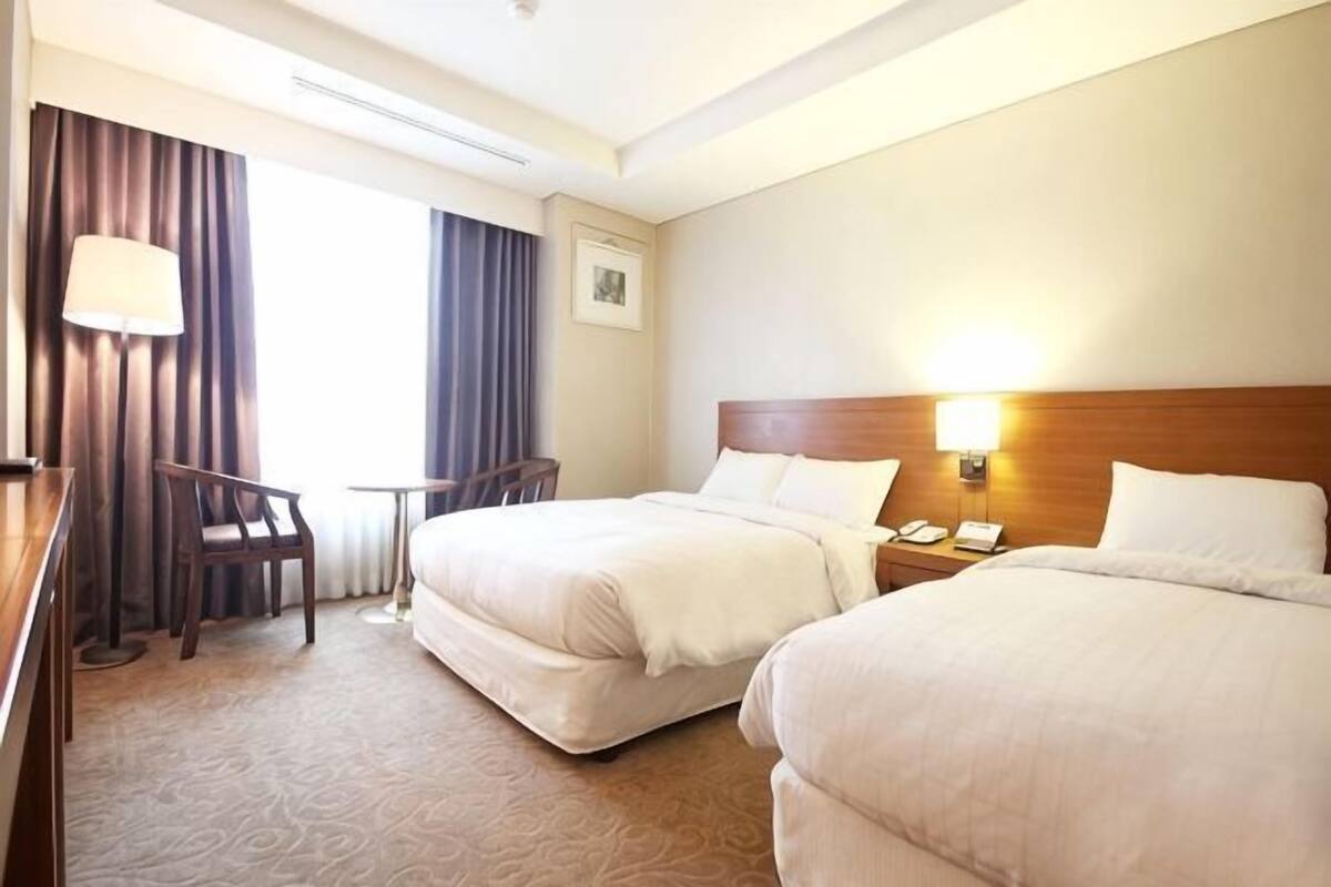A hotel room with two beds, a desk, a chair, a lamp, and a window with curtains.