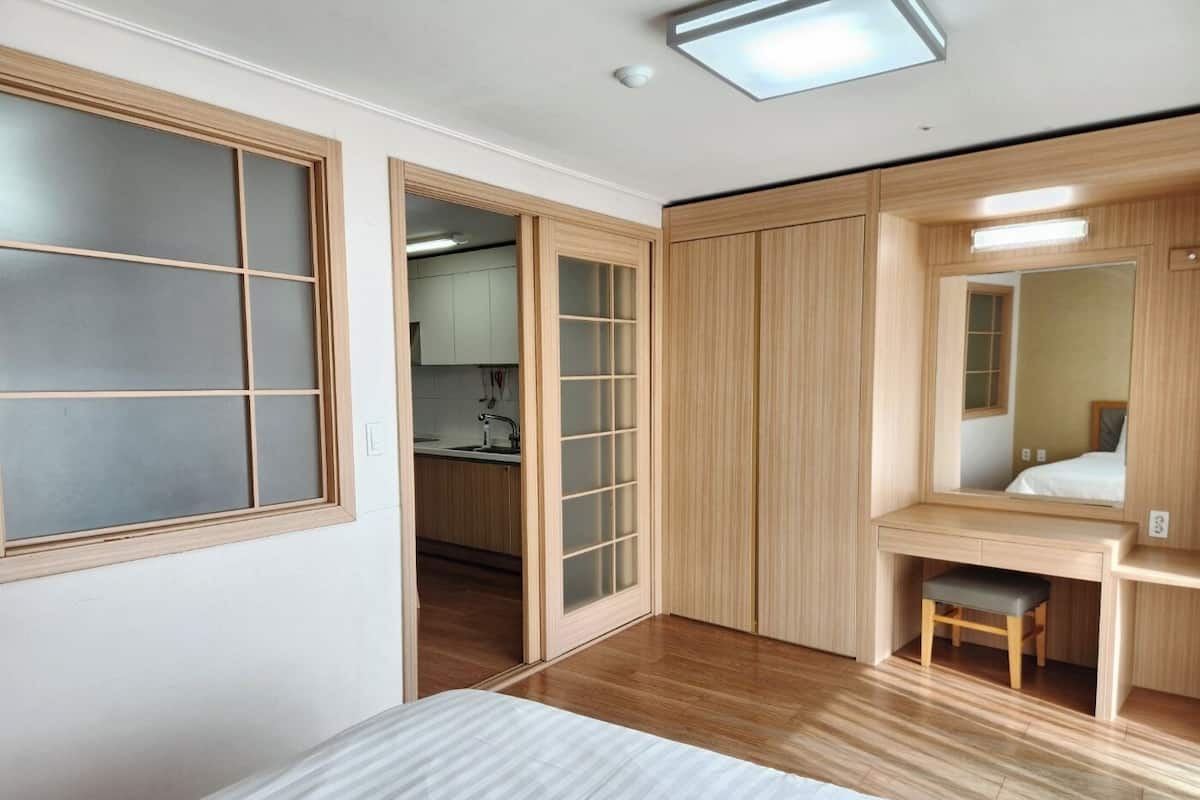A bedroom with a bed, a desk, a chair, a mirror, and a sliding door leading to a kitchen.