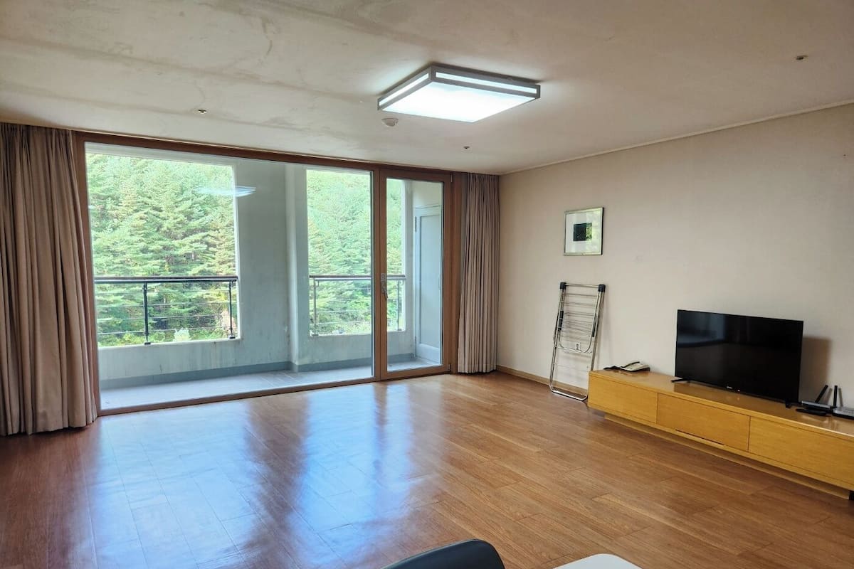 A spacious room with a large sliding glass door leading to a balcony, a flat-screen TV on a wooden cabinet, and a whiteboard on the wall.
