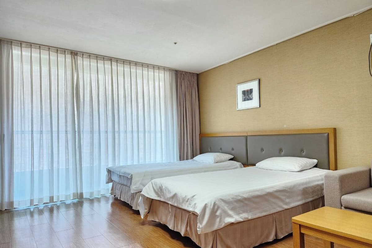 A hotel room with two beds, a sofa, a small table, a ceiling light, and a window with sheer curtains.