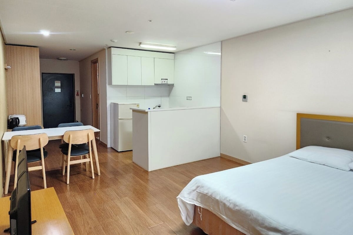 A compact hotel room with a bed, a desk with chairs, a kitchenette, and a door leading to another room.