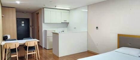 A compact hotel room with a bed, a desk with chairs, a kitchenette, and a door leading to another room.