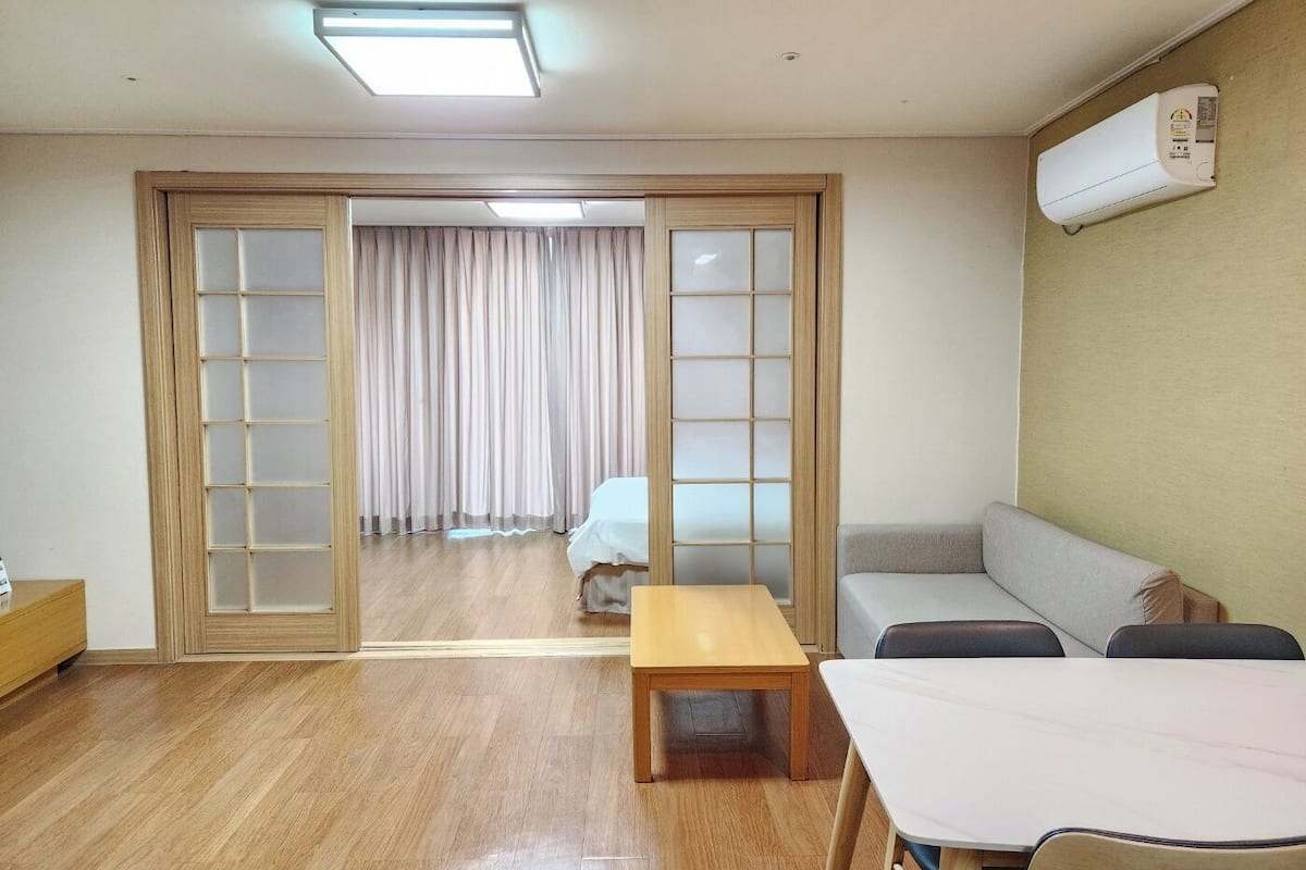 A room with a wooden floor, a sofa, a table, and a sliding door leading to a balcony.