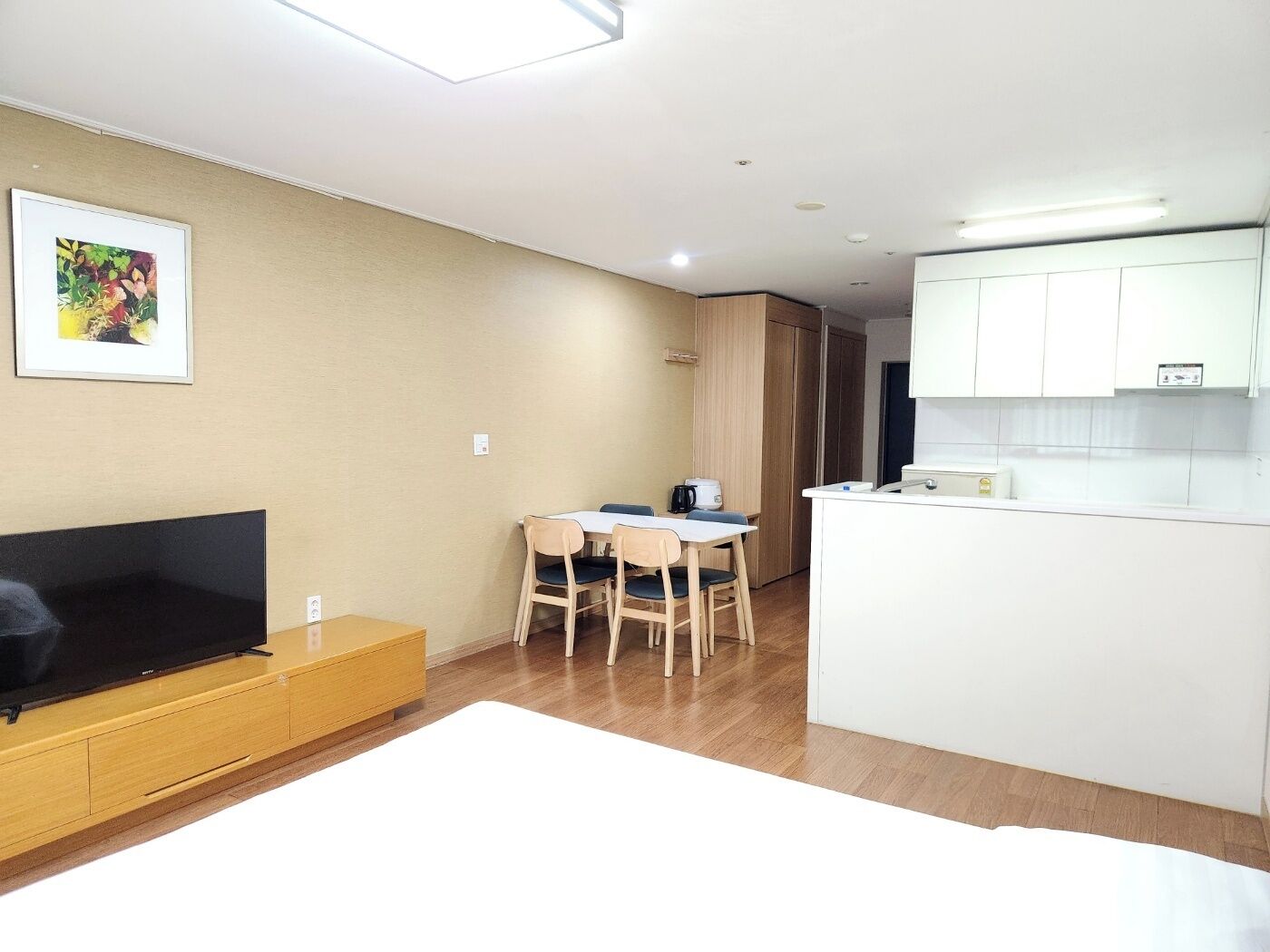 Una sala de estar moderna con televisor de pantalla plana, un comedor con mesa y sillas, y una kitchenette con armarios blancos.