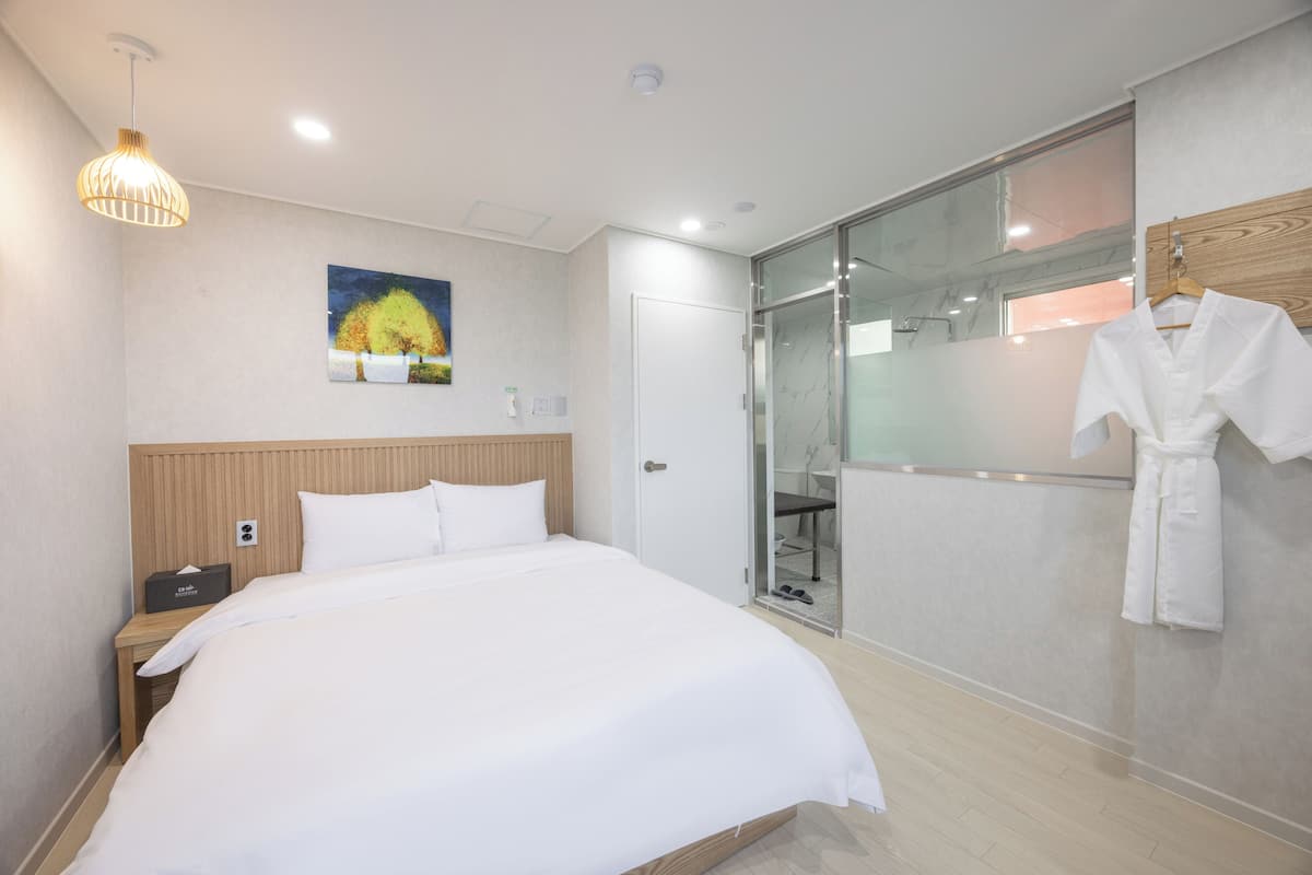 A modern hotel room with a large bed, a bedside table, a robe hanging on the wall, and a glass shower enclosure.