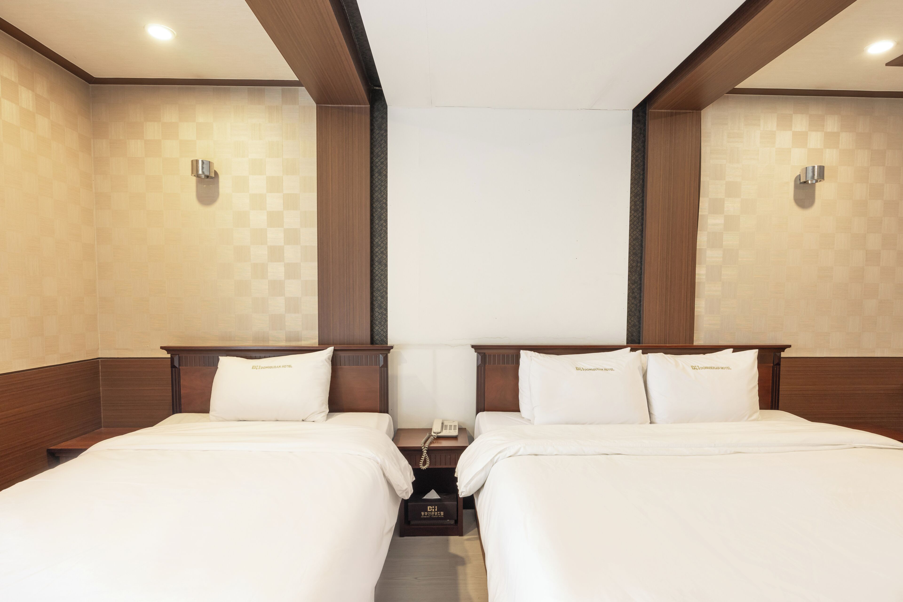 A hotel room with two beds, wooden headboards, a bedside table with a phone, and a wall-mounted light fixture.